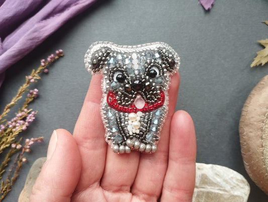 A hand holding a small, intricately beaded dog-shaped brooch against a gray background.