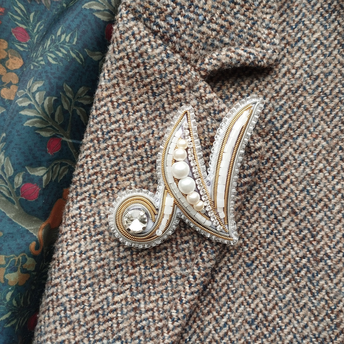 A close-up image of a decorative brooch or pin with a floral pattern and a crystal or gemstone at the center, attached to a tweed or woolen fabric.
