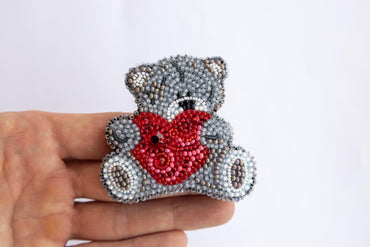 a hand holding a beaded teddy bear with a heart