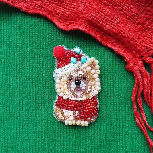 A beaded Christmas ornament in the shape of a dog wearing a Santa hat and scarf, placed on a green background with a red fringe.