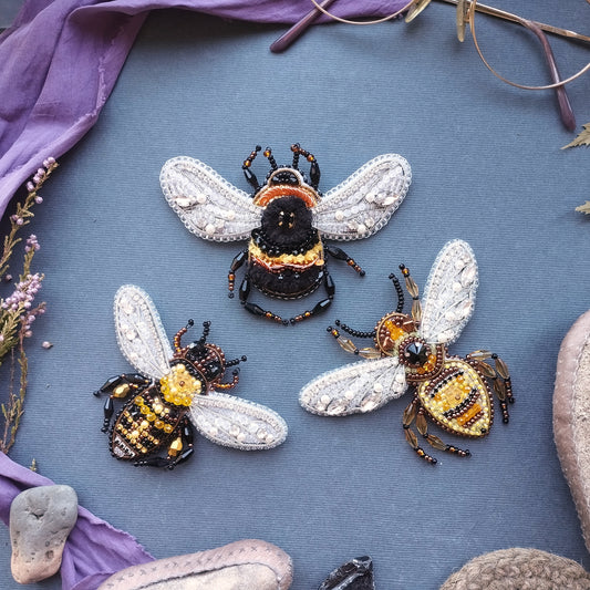 three intricately designed beaded bee brooches arranged on a blue background.