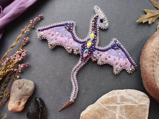 a decorative dragon-shaped object with intricate details and embellishments, including gemstones and beads, placed on a gray background alongside various natural elements such as rocks, flowers, and leaves.