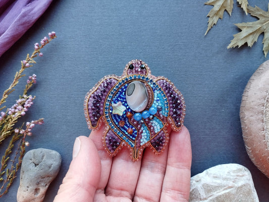 A hand holding a colorful, intricately beaded turtle brooch against a blue background.