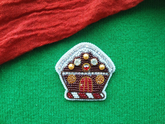 A decorative beaded house pin is displayed on a green fabric surface, with a red fabric item partially visible in the background.