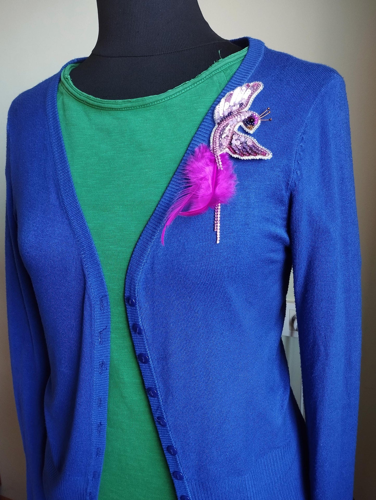 A mannequin wearing a blue cardigan with a green top underneath, and a pink feather brooch on the left side of the chest.
