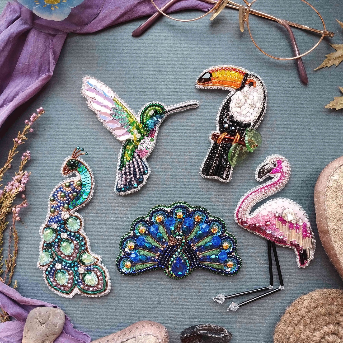 a collection of colorful, intricately designed beaded or embroidered animal pins or brooches, including a hummingbird, a peacock, a flamingo, and a parrot, all set against a purple background.