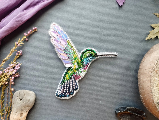 a colorful, intricately designed hummingbird brooch or pin, surrounded by various natural elements such as flowers, leaves, and rocks.