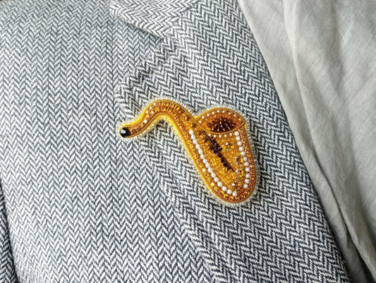 A close-up image of a gold and beaded brooch shaped like a saxophone, placed on a gray and white checkered fabric.