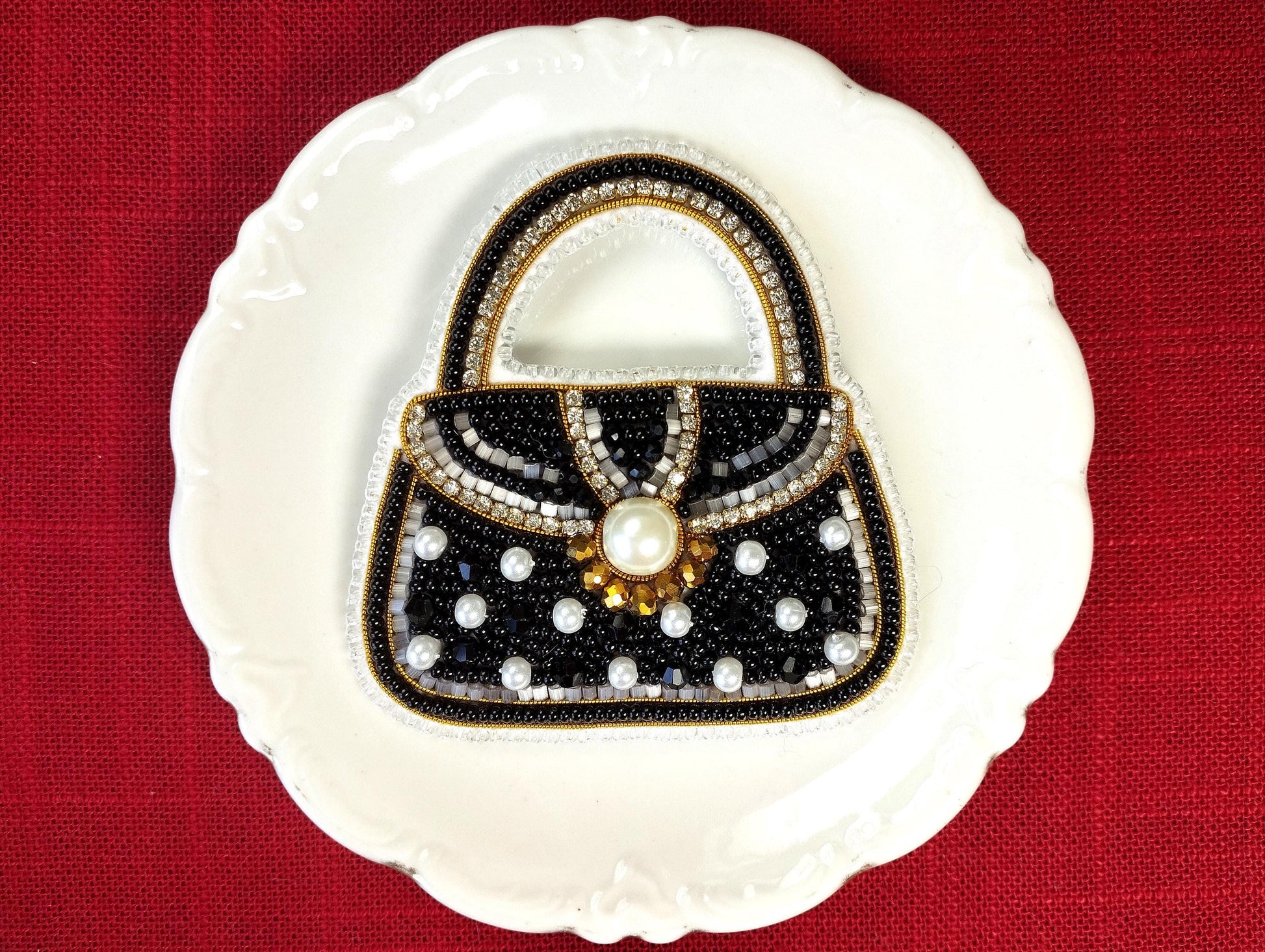 A white plate with a black and gold handbag-shaped design made of beads and pearls, placed on a red background.
