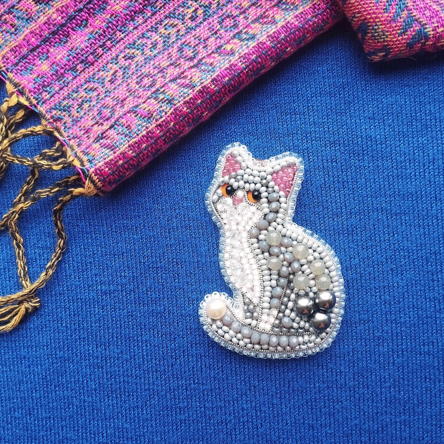 A beaded cat brooch is displayed on a blue fabric surface, with a pink and purple patterned fabric visible in the background.