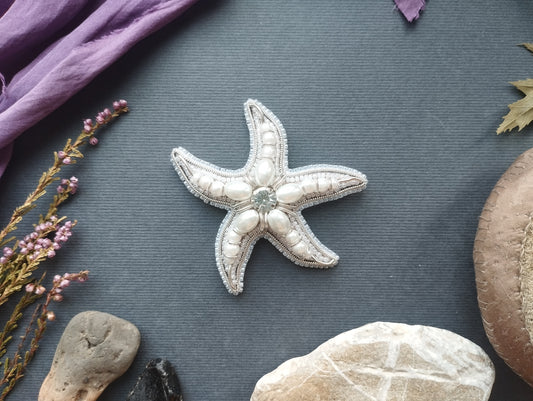 a close-up of a white starfish with a blue gemstone on it, surrounded by various natural elements such as rocks, leaves, and flowers on a gray background.