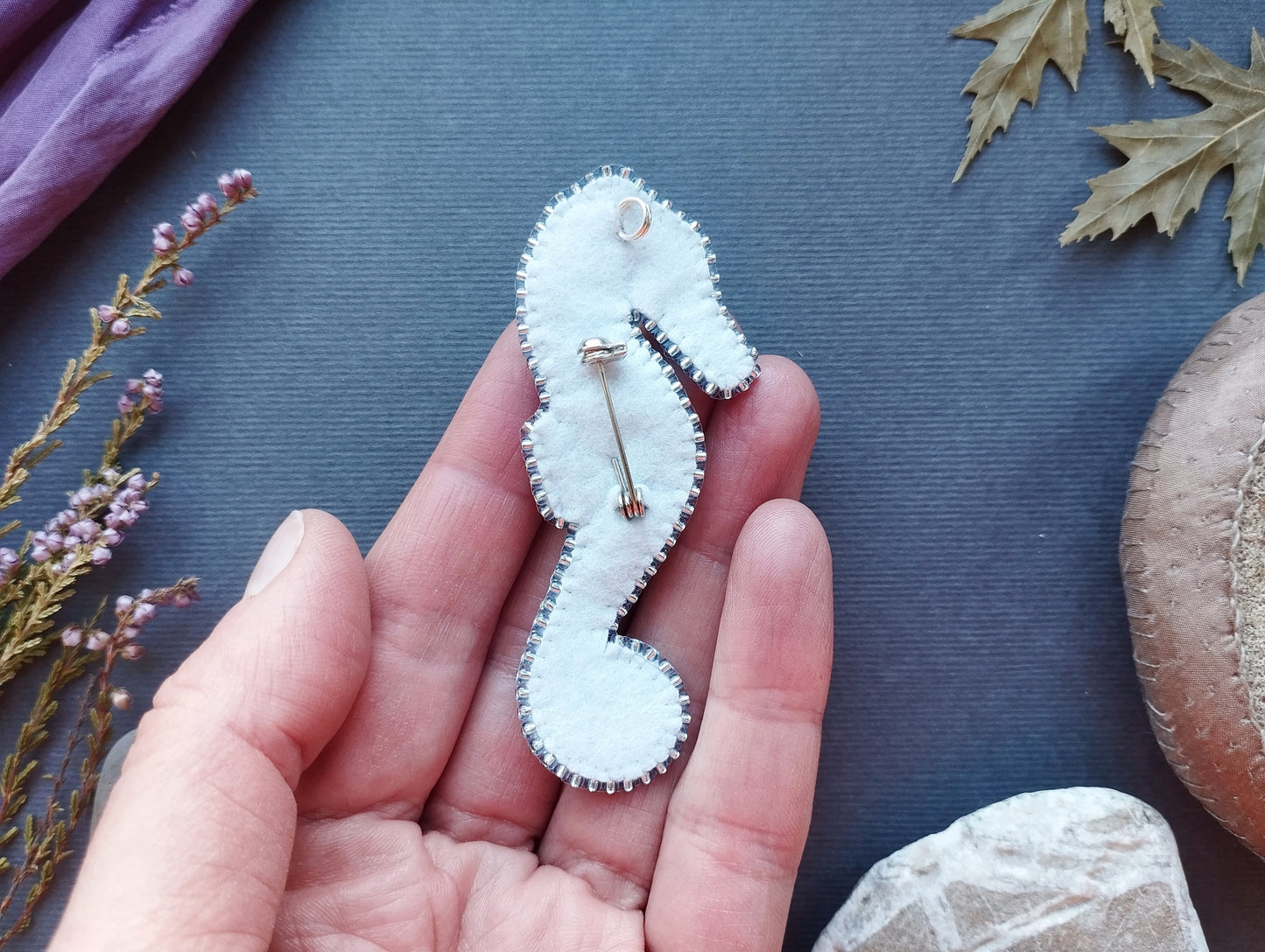 A hand holding a white felt seahorse pin against a blue background with some leaves and flowers.