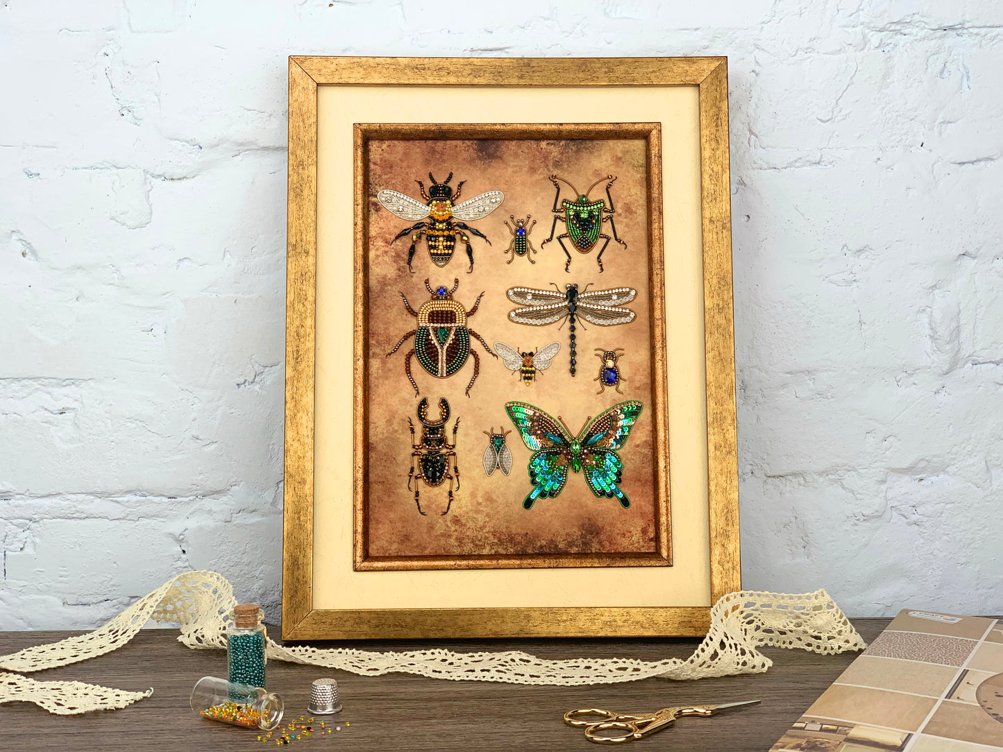 a framed collage of various insects, including bees, butterflies, and dragonflies, set against a brown background. The collage is displayed on a wooden surface, with a pair of scissors and some beads visible in the foreground.