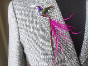 A close-up of a person wearing a gray tweed jacket with a colorful bird brooch on the left side of the jacket.
