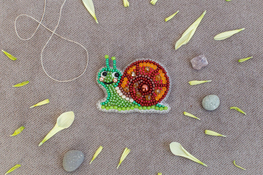 A colorful bead necklace depicting a snail is displayed on a beige surface, surrounded by scattered flower petals and small rocks.