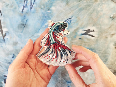 A hand holding a decorative beaded object resembling a fish or sea creature against a blue and white background.