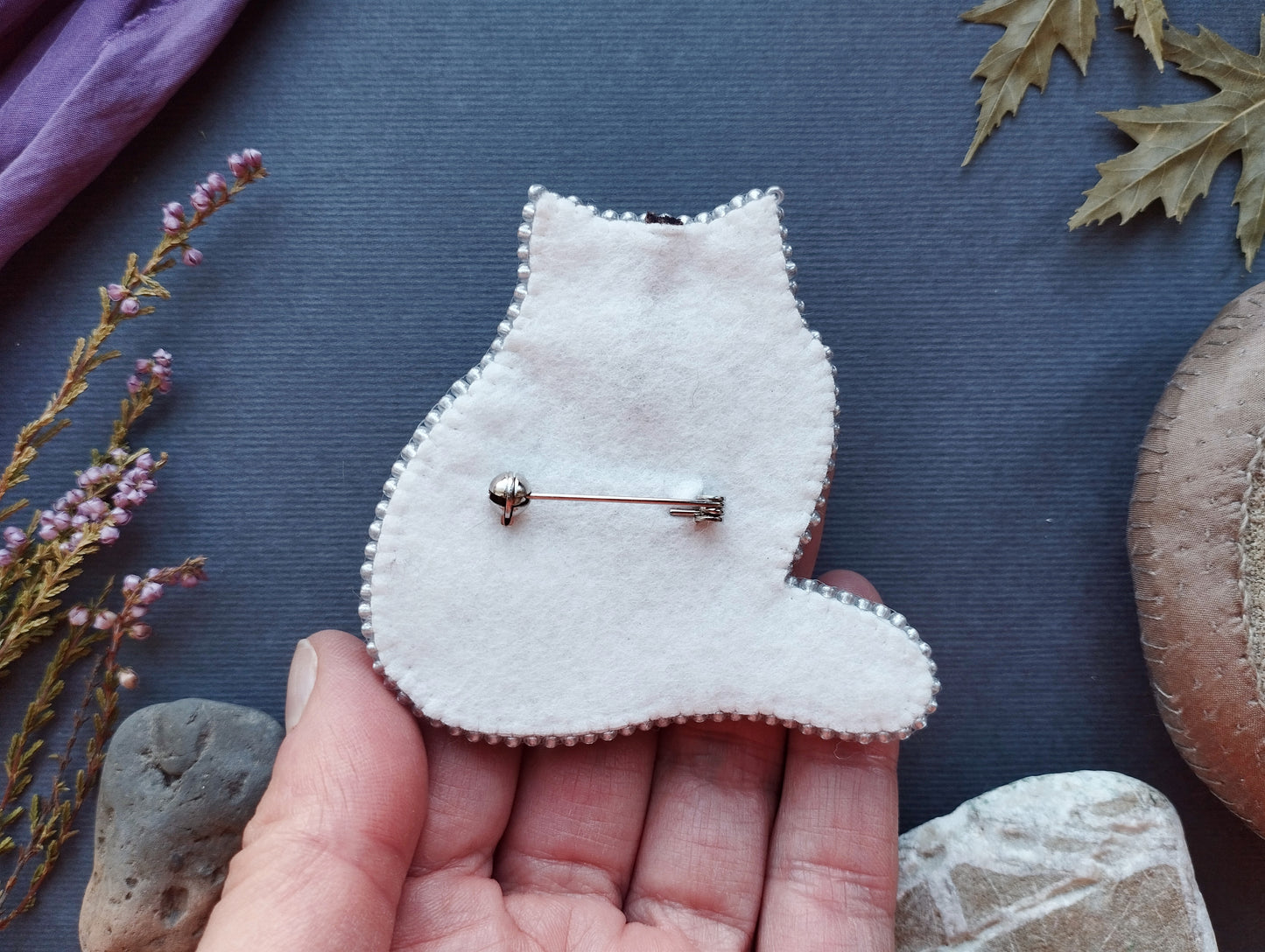 A hand holding a white felt cat pin against a dark background with various objects, including leaves and rocks.