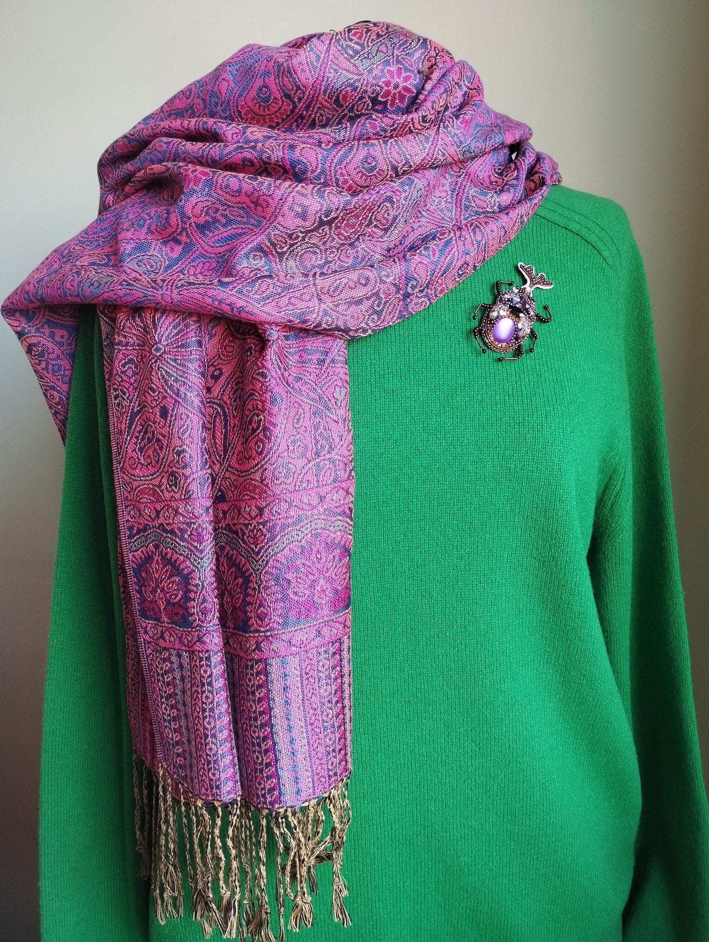 A mannequin is wearing a green sweater and a pink and purple patterned scarf with fringe, and a brooch is pinned to the sweater.