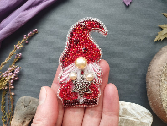 A hand holding a red and silver ornament with a gnome-like design, adorned with beads and pearls.