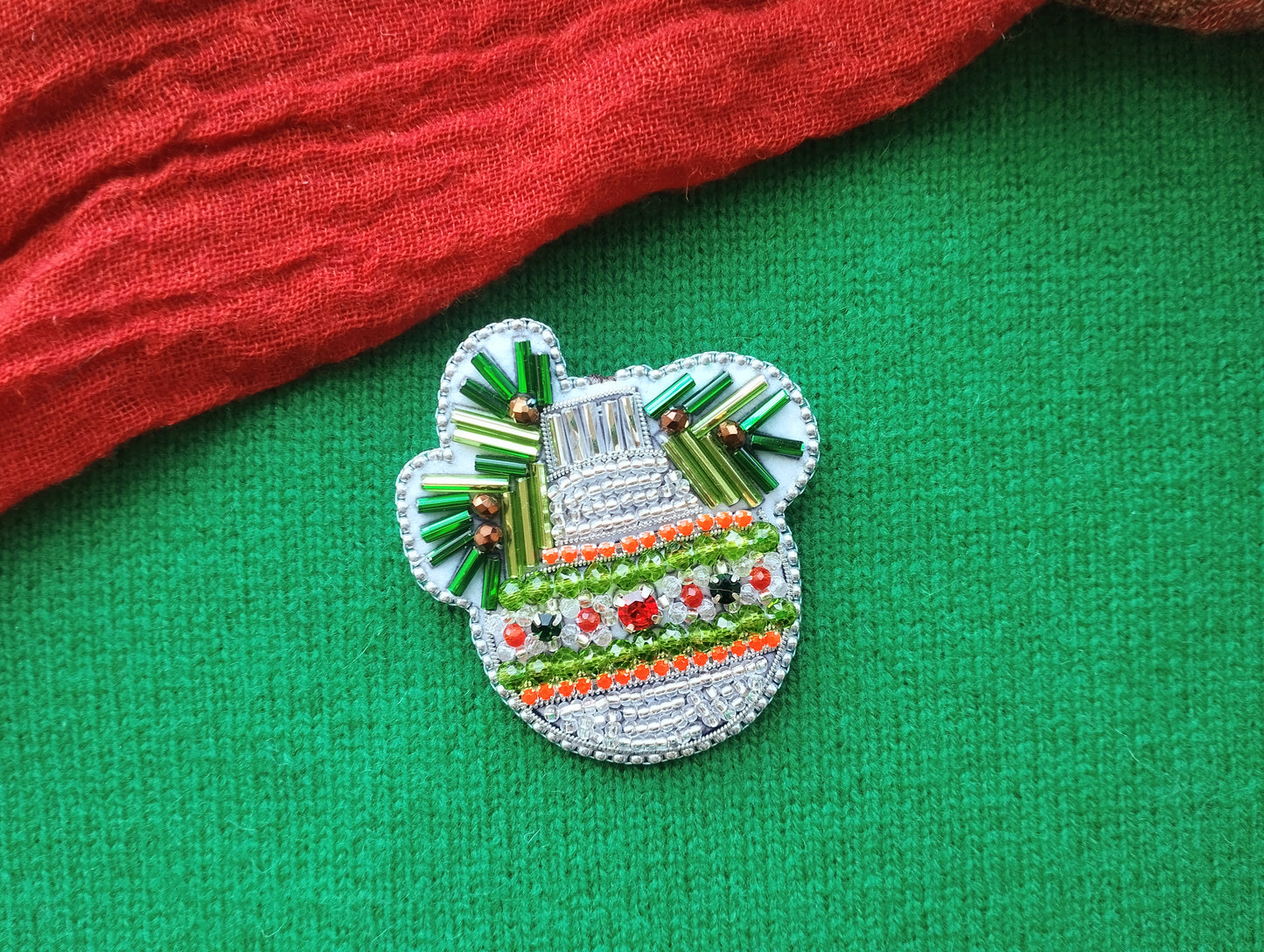 A decorative Christmas ornament with a silver and green design, featuring a bell-shaped motif and colorful beads, is displayed on a green fabric background.