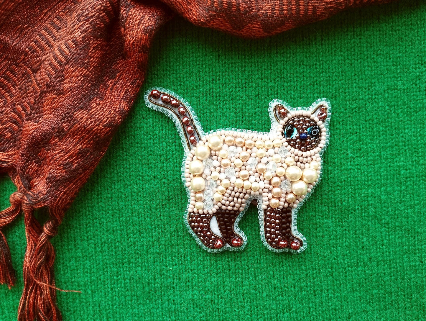 a decorative cat brooch made of beads and pearls, set against a green background.