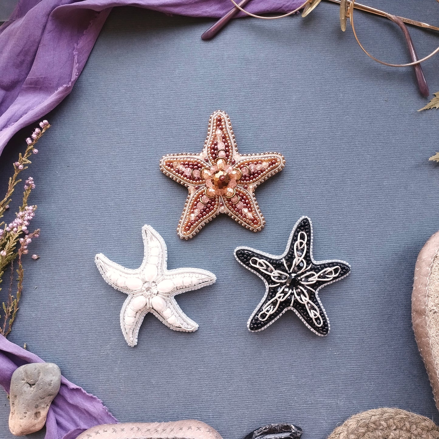 three starfish or sea starlets, each with a unique design, arranged on a blue background. The starfish on the left is white, the one in the middle is brown, and the one on the right is black. The starfish on the left appears to be the largest, while the black starfish on the right is the smallest. The starfish on the right is positioned slightly higher than the other two, creating a sense of depth in the image.