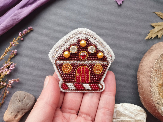 A hand holding a small, intricately beaded and embroidered object resembling a gingerbread house or a similar decorative piece.