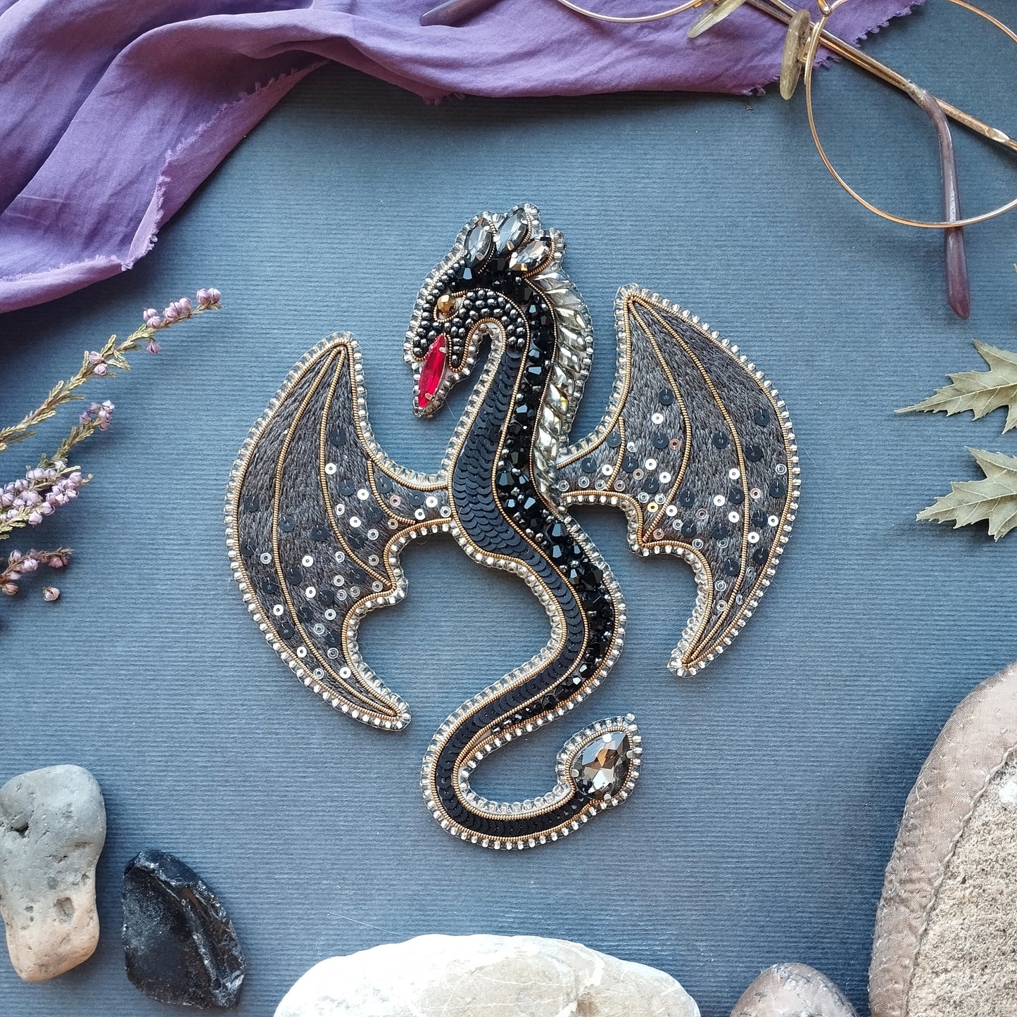 a detailed embroidered dragon brooch with intricate patterns and sparkling gemstones, surrounded by various natural elements such as leaves, rocks, and a purple fabric.
