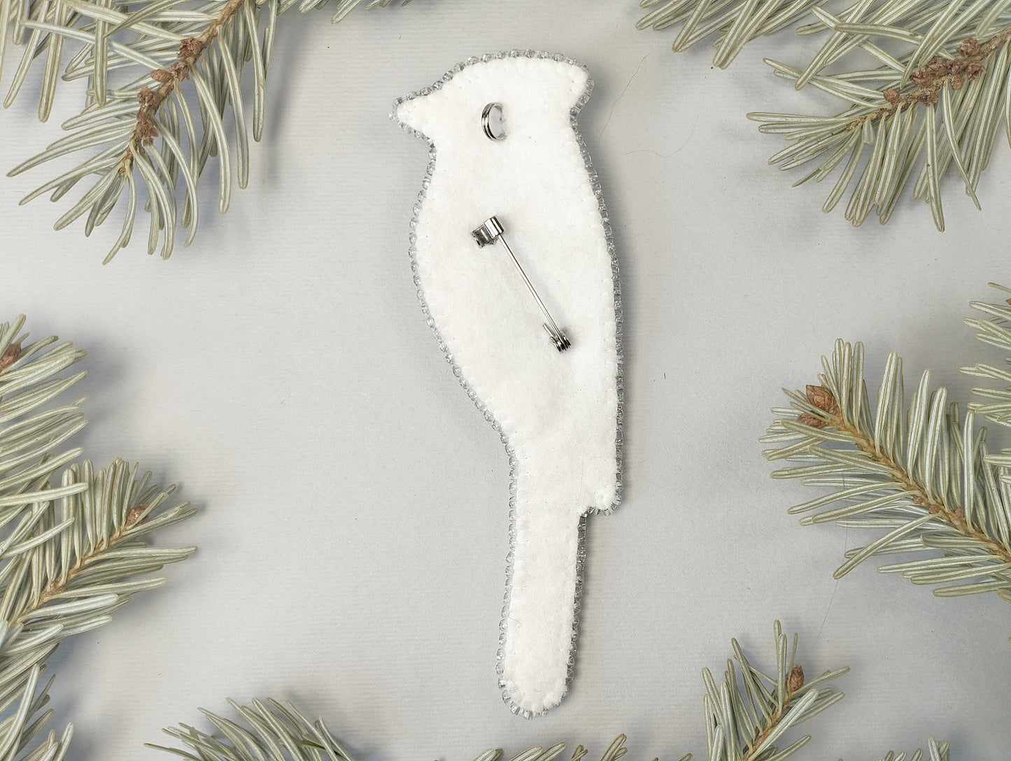 A white bird-shaped ornament with a silver pin attached to it, surrounded by pine needles on a light gray background.