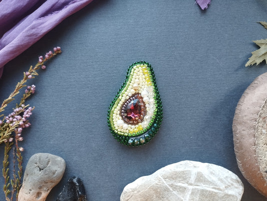 a decorative piece of jewelry, specifically a brooch, with a vibrant design of green, yellow, and red beads arranged to resemble an avocado. The brooch is placed on a gray surface, surrounded by various natural elements such as leaves, rocks, and flowers.
