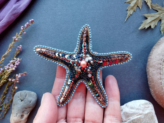 A hand holding a starfish adorned with colorful beads and sequins, surrounded by various natural elements such as leaves, rocks, and flowers.