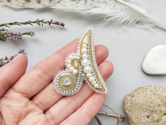 A hand holding a decorative piece of jewelry, which appears to be a brooch or pin, with a combination of pearls, beads, and a gemstone. The background includes feathers, rocks, and flowers, creating a natural and artistic setting.