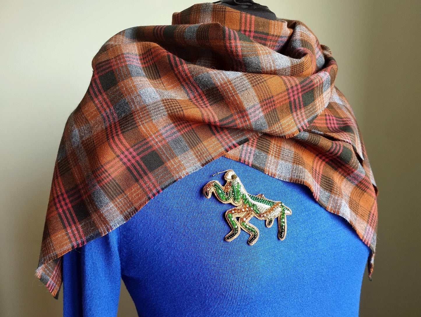 A plaid scarf is draped over a blue sweater, with a green and gold brooch shaped like a horse pinned to the sweater.