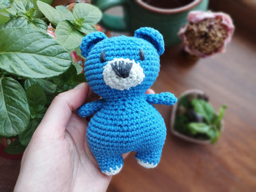 a crocheted blue teddy bear sitting next to a potted plant