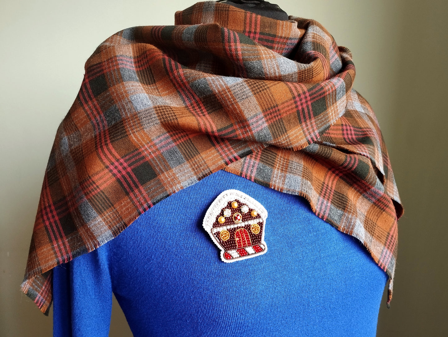 A plaid scarf is draped over a blue sweater, with a small crochet patch featuring a gingerbread house design on the sweater.