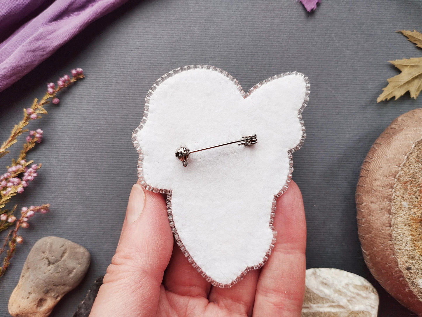 A hand holding a white felt pin or badge against a gray background, with various natural elements such as leaves, flowers, and rocks scattered around.
