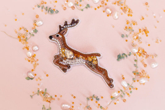 a decorative deer embroidered with jewels and surrounded by various beads and pearls on a pink background.