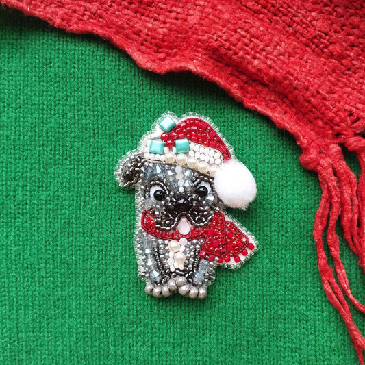 A decorative beaded brooch in the shape of a dog wearing a Santa hat and scarf, placed on a green fabric background with a red fringe.