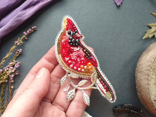 A hand holding a decorative bird brooch with sequins and leaves on a gray background.