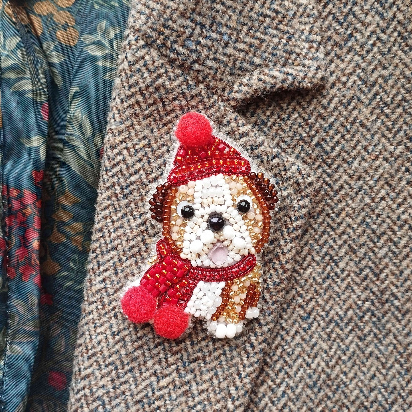 A close-up of a small, intricately beaded dog-shaped brooch or pin, wearing a red hat and scarf, attached to a tweed jacket.