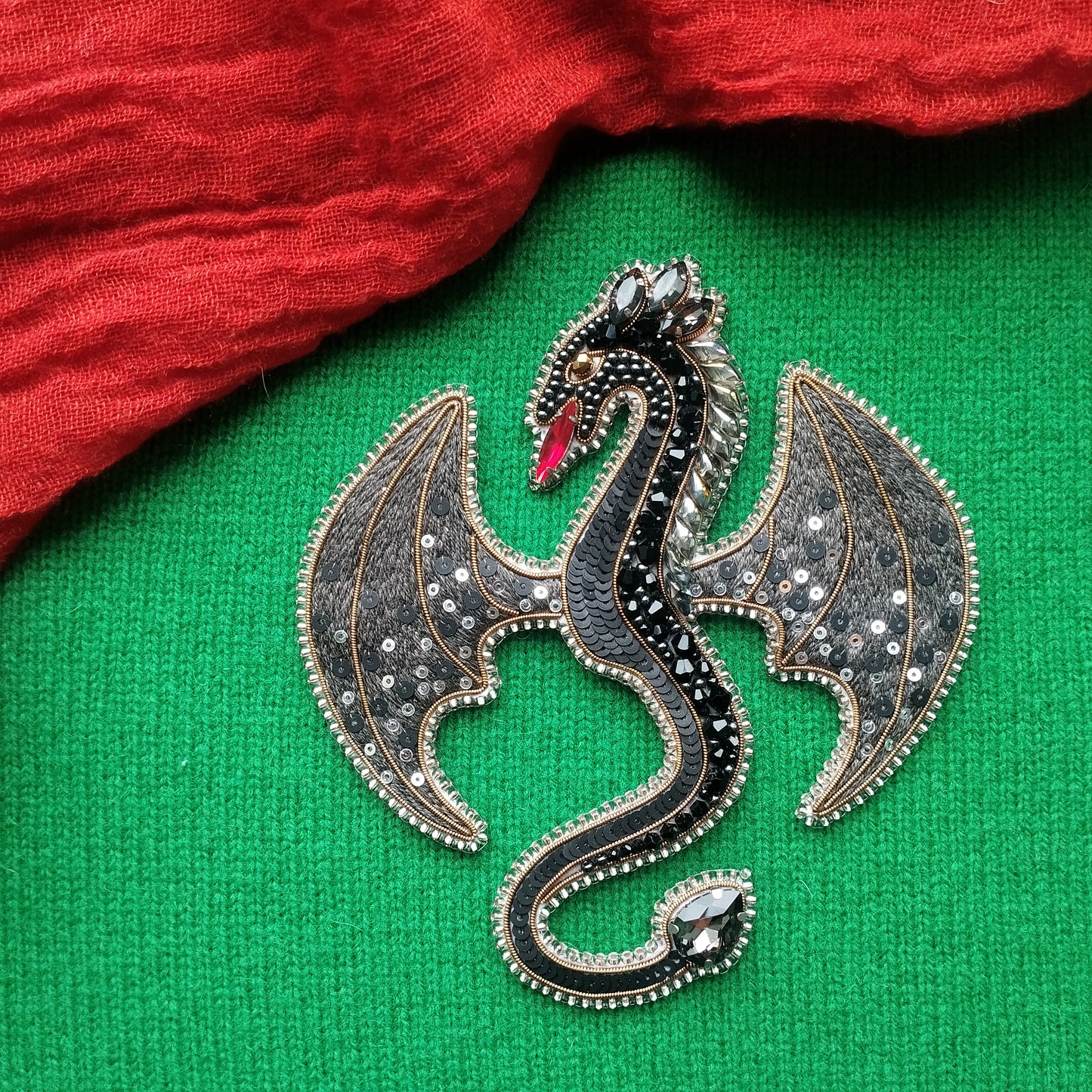 A decorative dragon brooch with a black and silver design, featuring a red eye and wings, is displayed on a green fabric background.