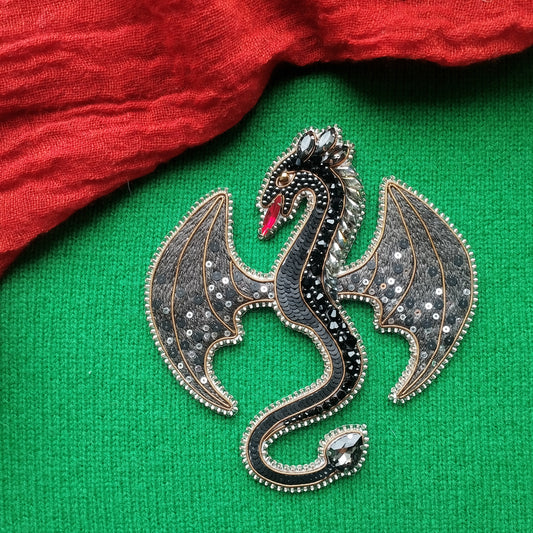 A decorative dragon brooch with a black and silver design, featuring a red eye and wings, is displayed on a green fabric background.