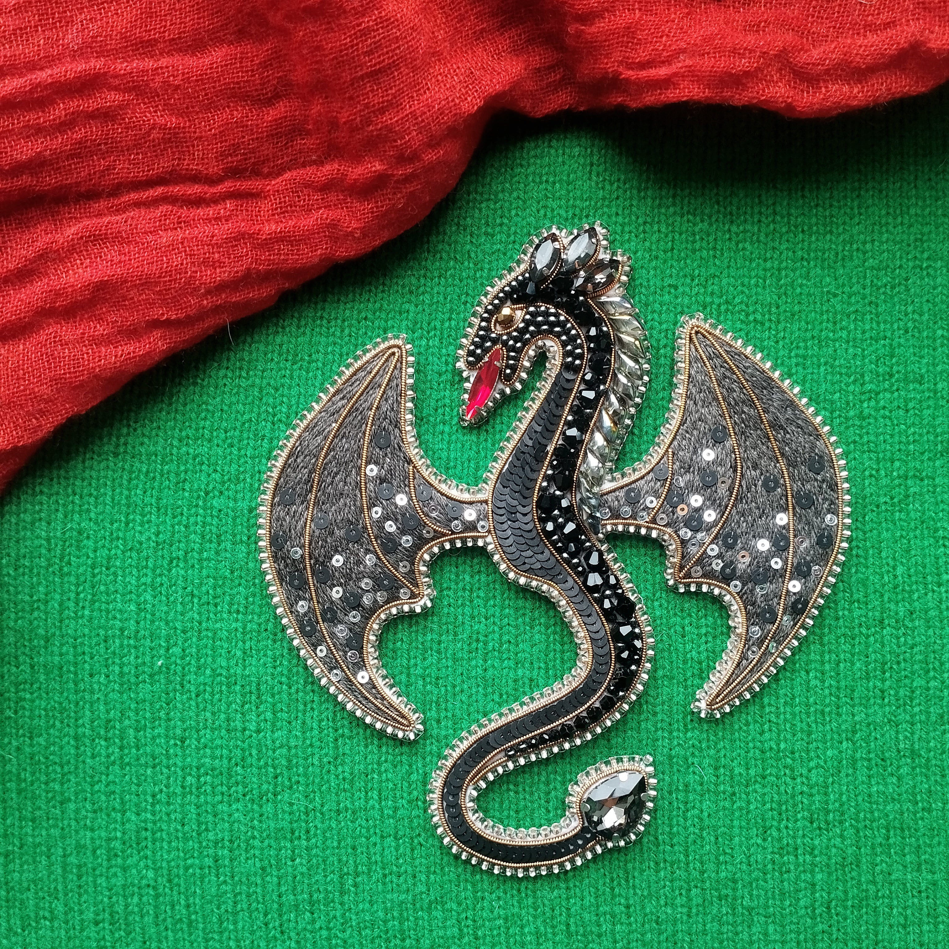 A decorative dragon brooch with a black and silver design, featuring a red eye and wings, is displayed on a green fabric background.
