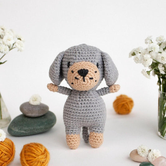A crocheted gray dog toy with a black nose and eyes is surrounded by various objects, including flowers, rocks, and balls of yarn, creating a whimsical and playful scene.