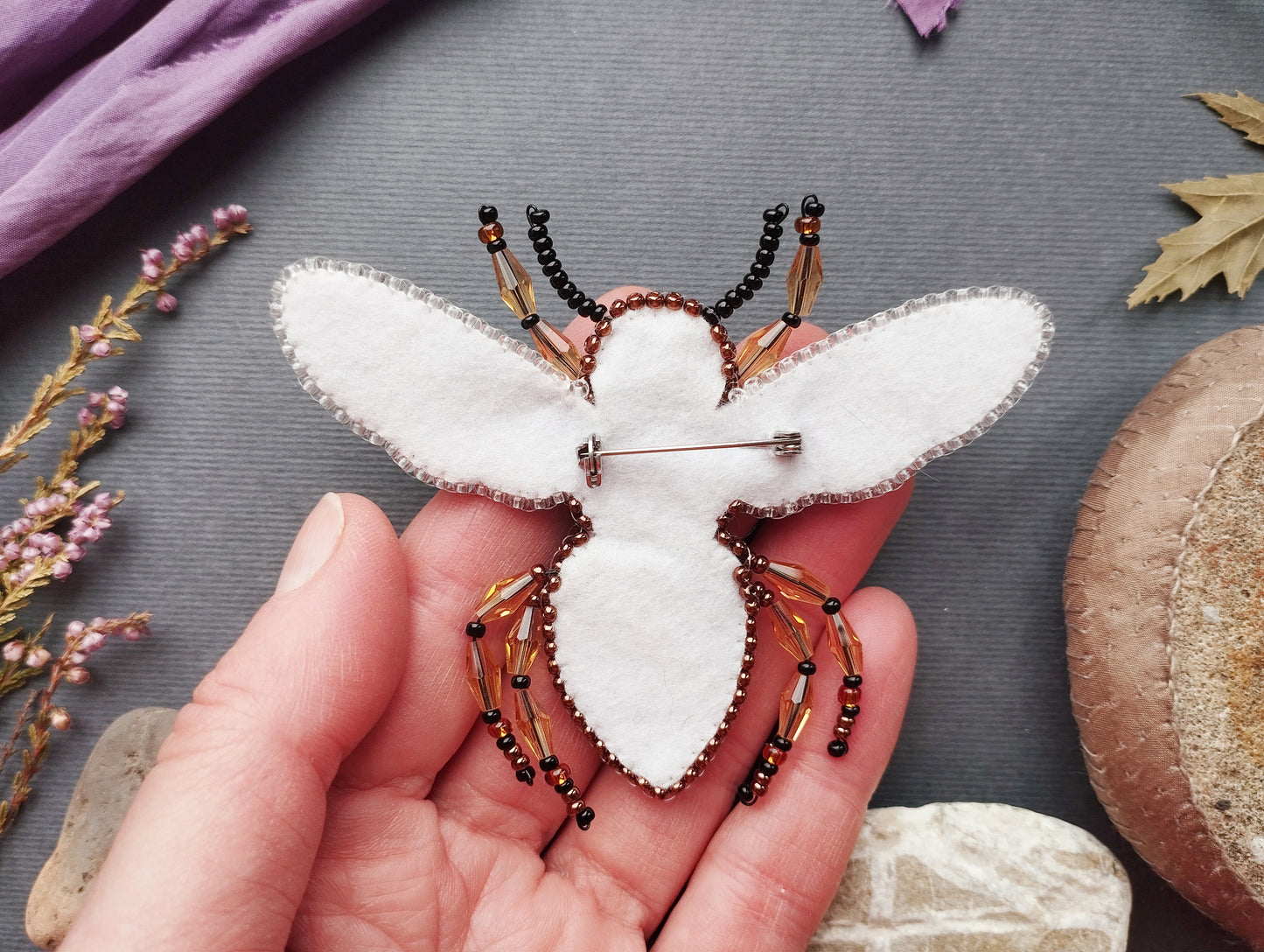 A hand holding a white felt insect pin adorned with beads, surrounded by various natural elements such as leaves and flowers.