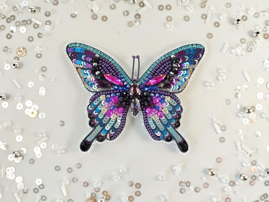 A colorful, sequined butterfly is the central focus of the image, surrounded by scattered sequin pieces on a light background.