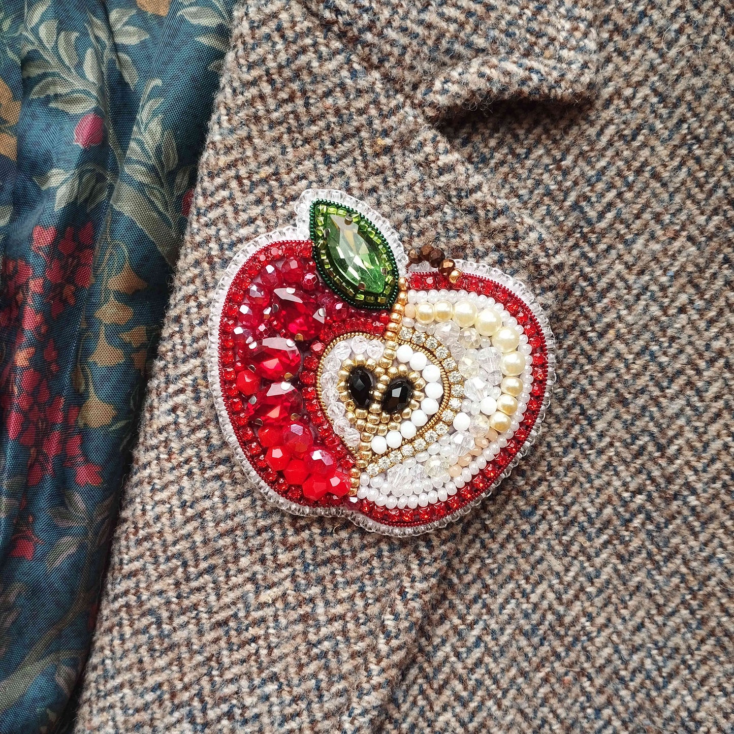 A close-up image of a decorative brooch in the shape of an apple, adorned with various colorful gemstones and beads.