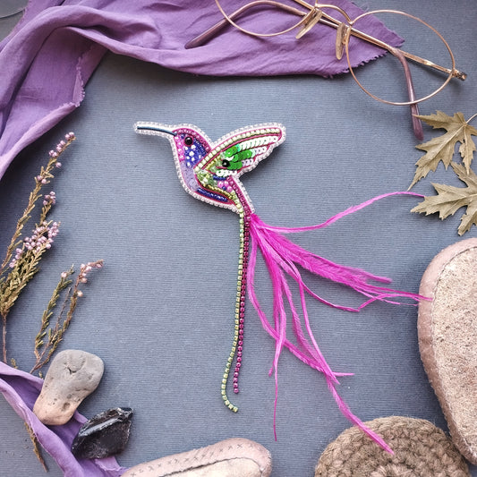 a colorful, hand-made hummingbird brooch with a long pink feather, surrounded by various natural elements such as leaves, flowers, and rocks.