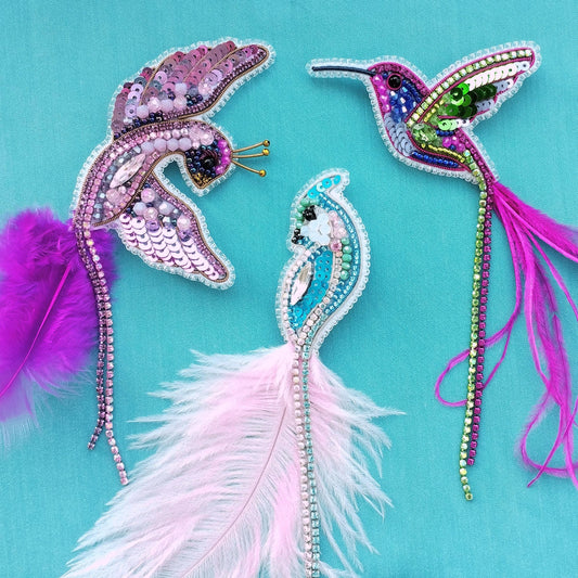 three colorful bird brooches with intricate beading and feathers, set against a teal background.