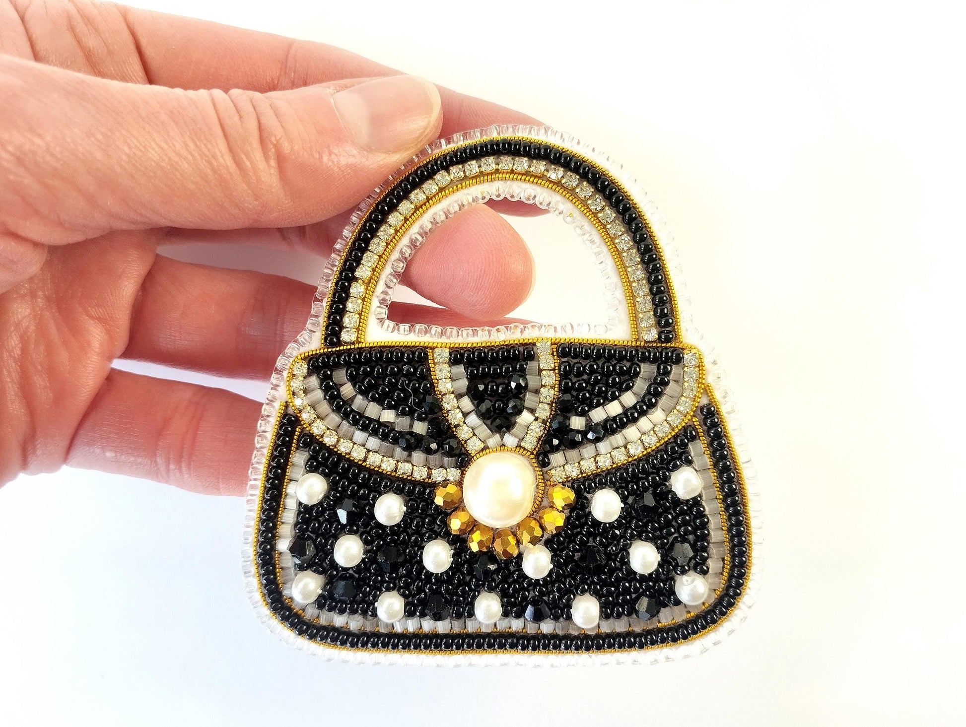 A hand holding a handbag-shaped embroidered piece with black, white, and gold beads and a pearl at the center.